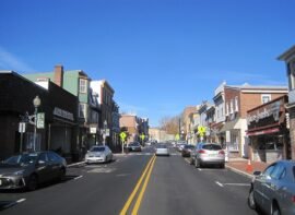 High Street in downtown Mount Holly, NJ, featuring historic buildings, local businesses, and parked cars along a two-lane road under a clear blue sky.