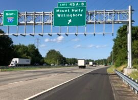 Highway exit sign for Mount Holly and Willingboro, NJ on Interstate 295, surrounded by green trees and traffic under a blue sky.